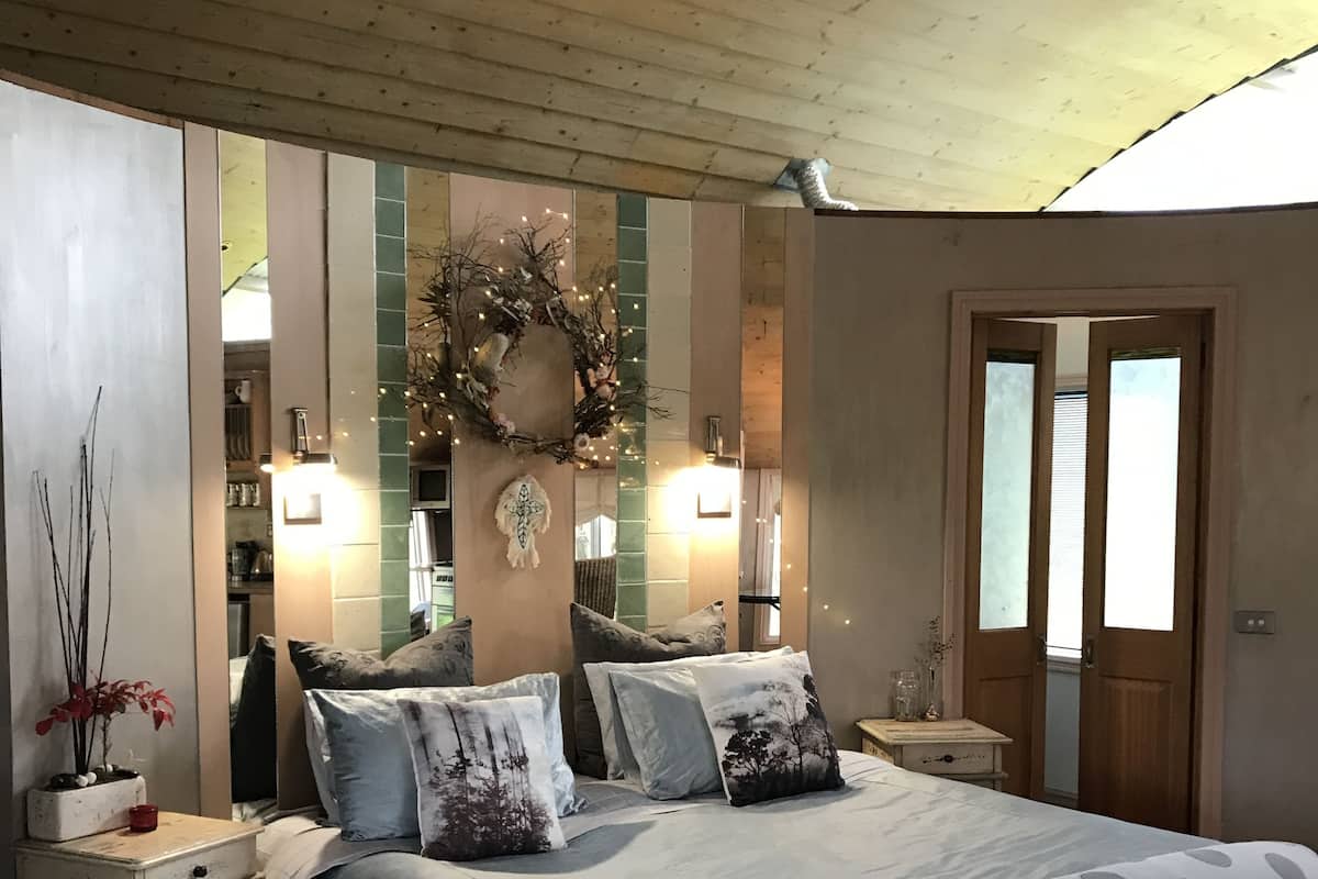 A bedroom with a bed, nightstands, a chair, a wooden ceiling, a door, and a mirror.