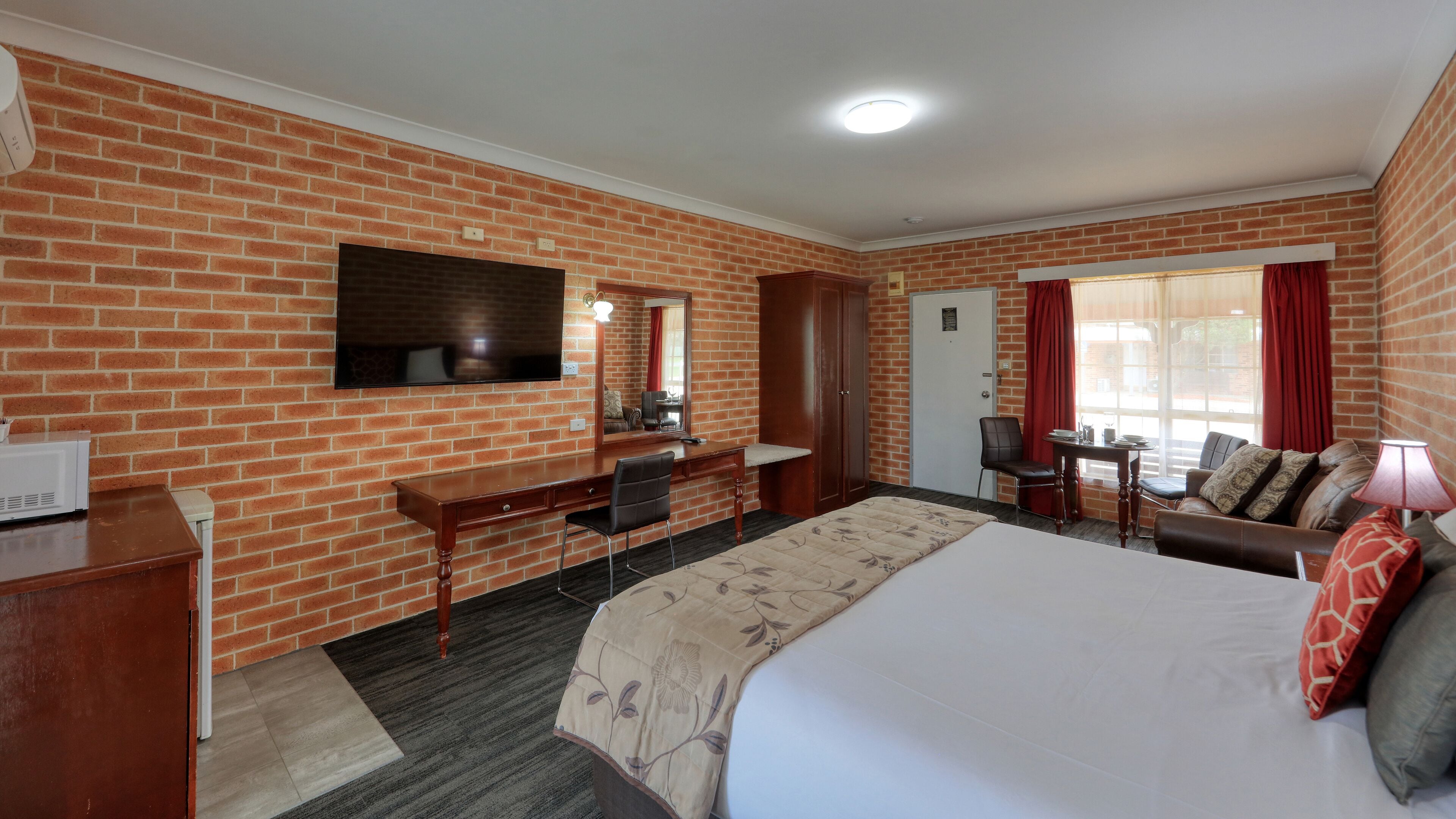 A hotel room with a brick wall, a bed, a desk with a chair, a TV mounted on the wall, a door, and a window with curtains.