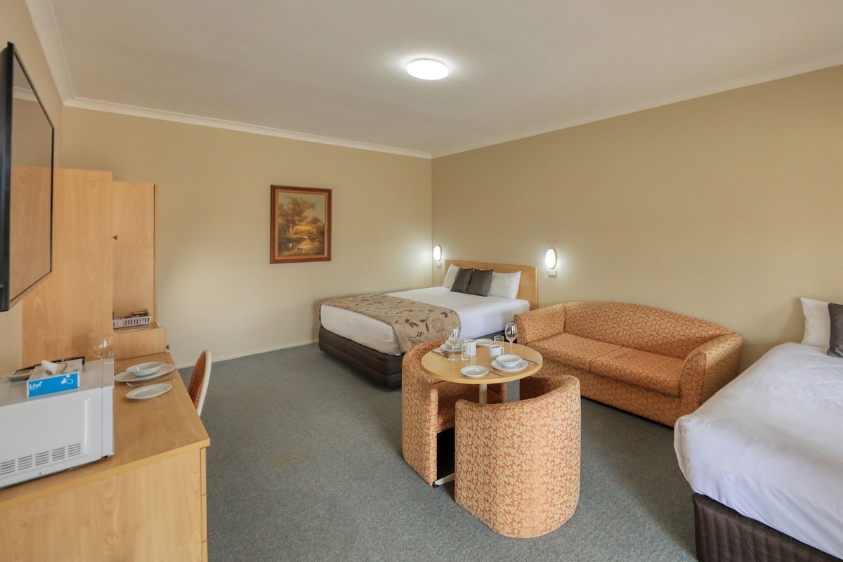 Habitación de hotel con cama, sofá, una pequeña mesa con vasos y platos, televisión y un cuadro en la pared.