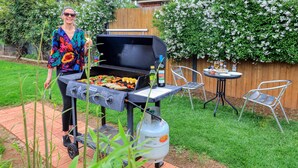 Un barbecue avec des légumes et des saucisses en train de cuire, une bouteille de gaz et une table avec des chaises installées à proximité.