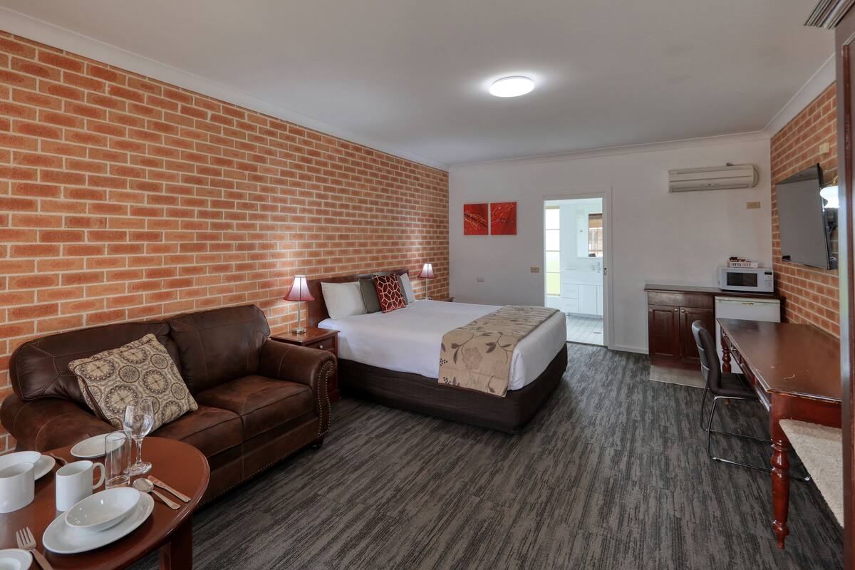 Una habitación de hotel con pared de ladrillo, una cama con colcha blanca, un sofá de cuero marrón y una mesa de comedor con platos, vasos y cubiertos.