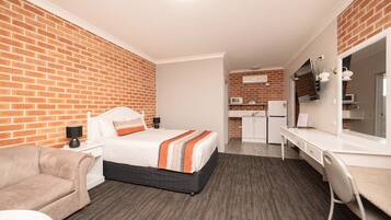 A hotel room with a brick wall, a bed with a striped blanket, a desk with a chair, a kitchenette, and a flat-screen TV.