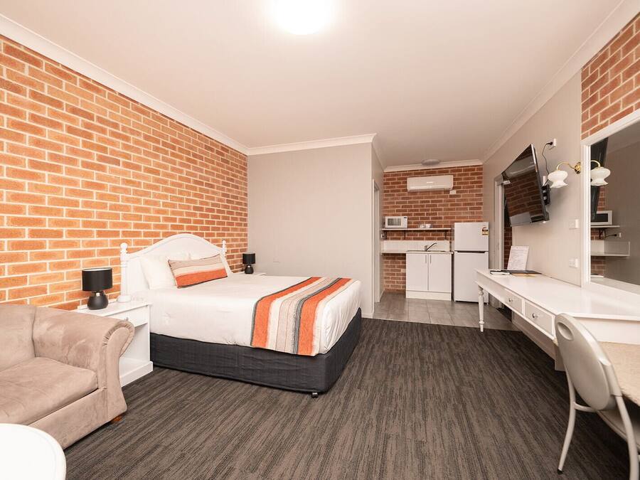 A hotel room with a brick wall, a bed with a striped blanket, a desk with a chair, a kitchenette, and a flat-screen TV.