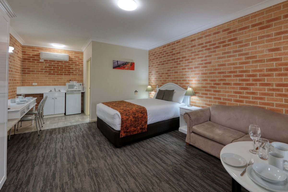 Habitación de hotel con cama, sofá, mesa de comedor con platos y vasos, kitchenette y pared de ladrillo.
