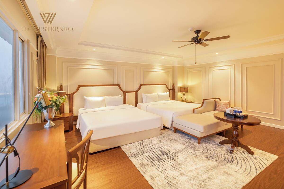 A hotel room with two beds, a desk, a chair, a small table, a ceiling fan, and a large window.