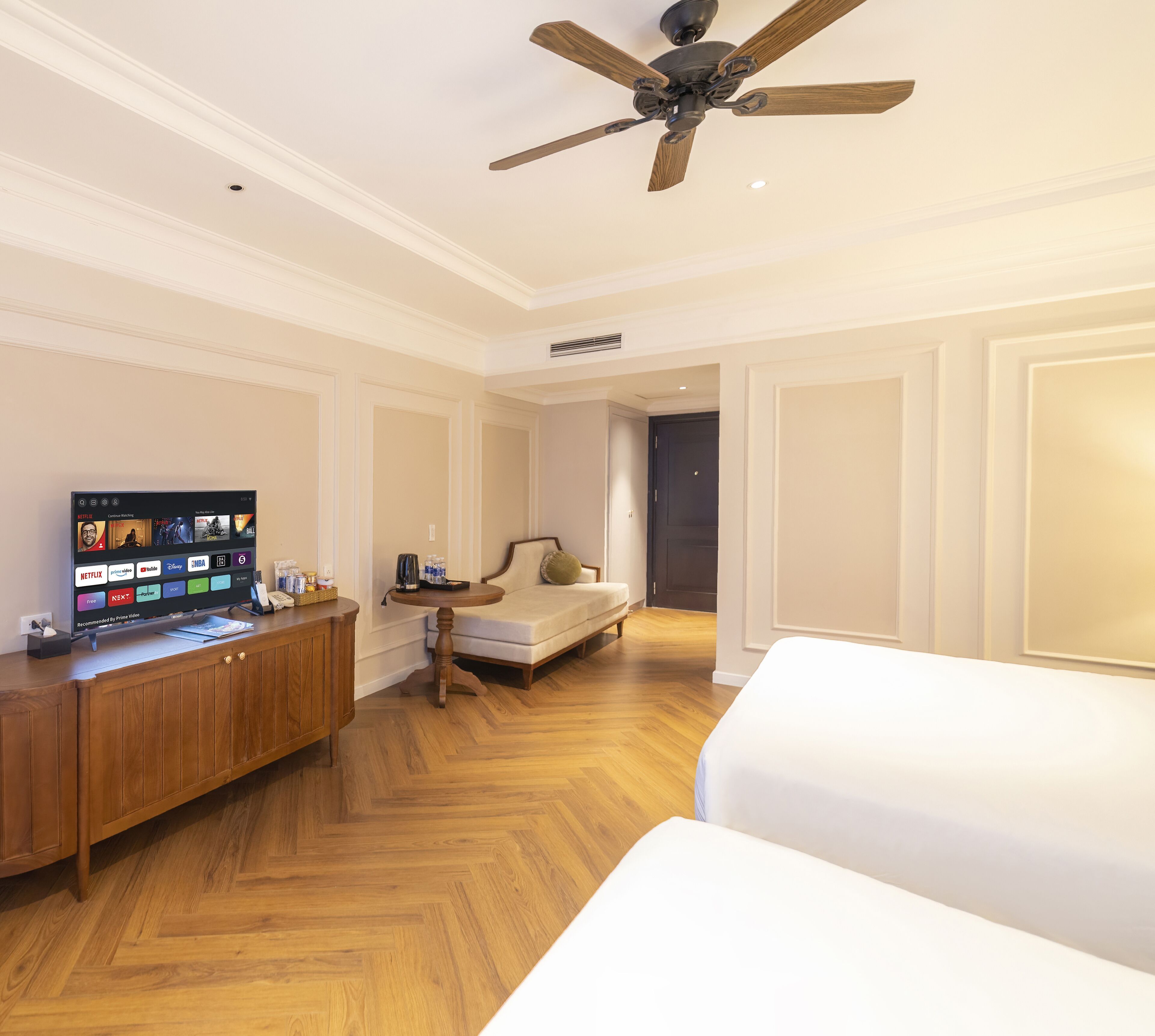 A hotel room with a wooden floor, a bed, a sofa, a TV on a wooden cabinet, and a ceiling fan.