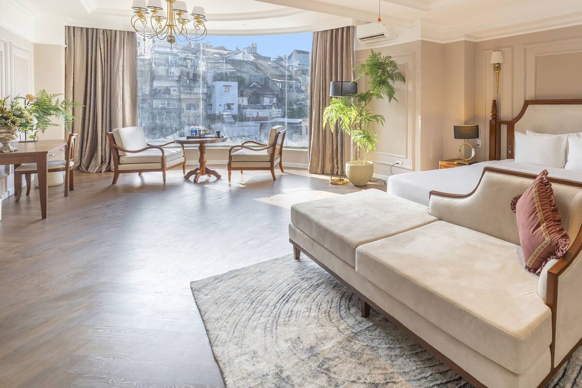 A hotel room with a large bed, a sofa, a dining table with chairs, and a view of the city.