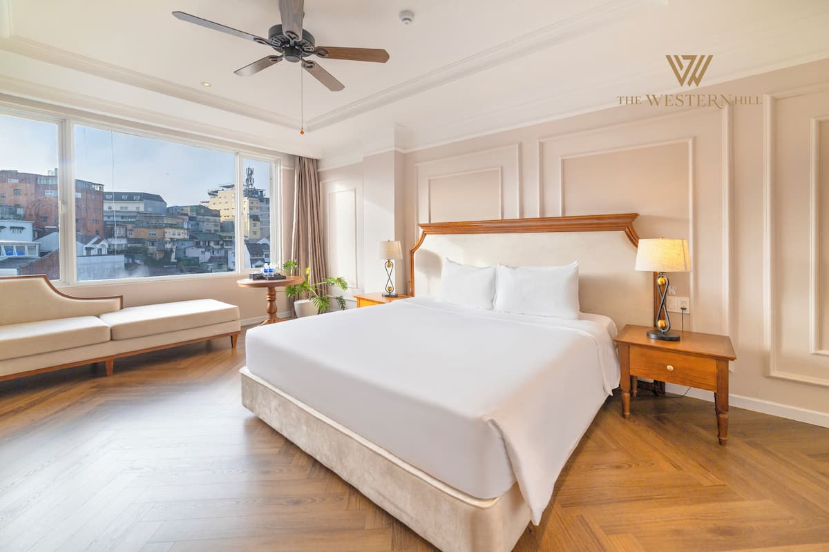 A hotel room with a large bed, a sofa, a nightstand with a lamp, and a view of the city through large windows.