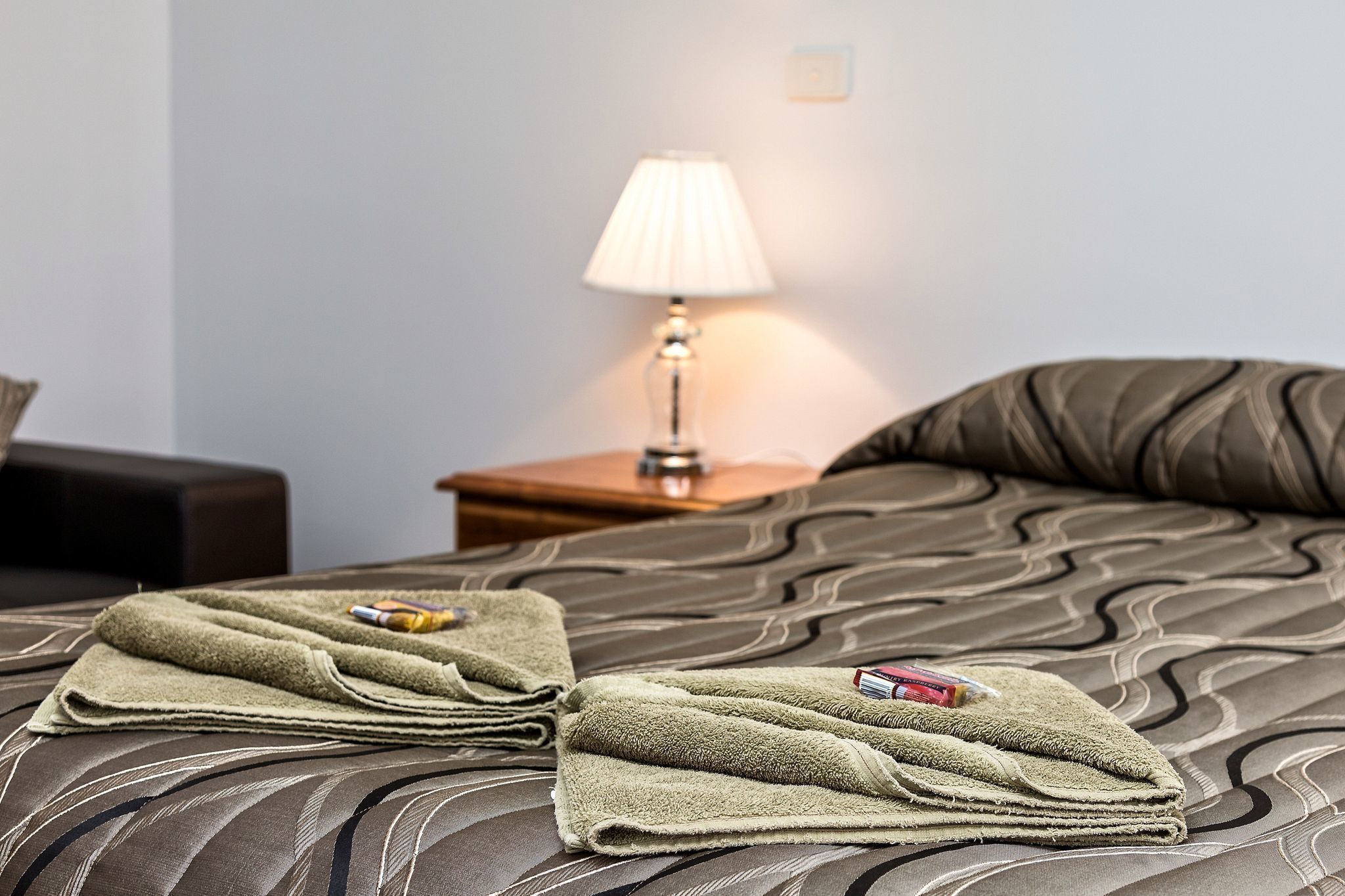 A neatly made bed with a patterned bedspread, a folded green towel, and a bottle of lotion on the bed.