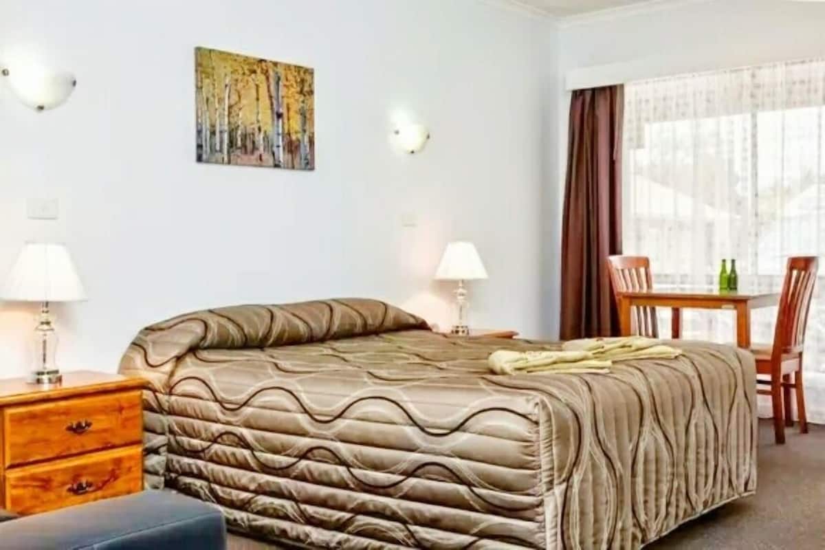 A hotel room with a bed, bedside tables, a chair, a table, and a painting on the wall.