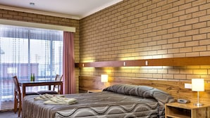A hotel room with a brick-patterned wall, a bed with a quilted cover, a bedside table with a lamp, and a dining area with a table and chairs. - Holbrook Town Centre Motor Inn (Holbrook)
