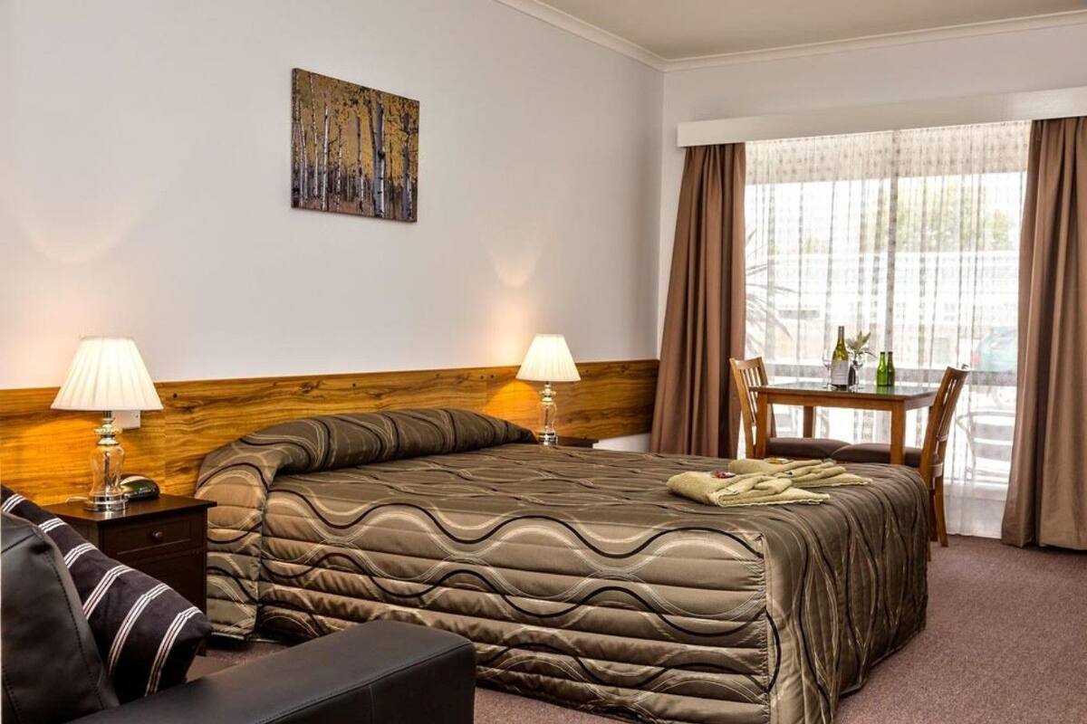 A hotel room with a large bed, two bedside tables with lamps, a wooden headboard, a dining table with chairs, and a window with curtains.