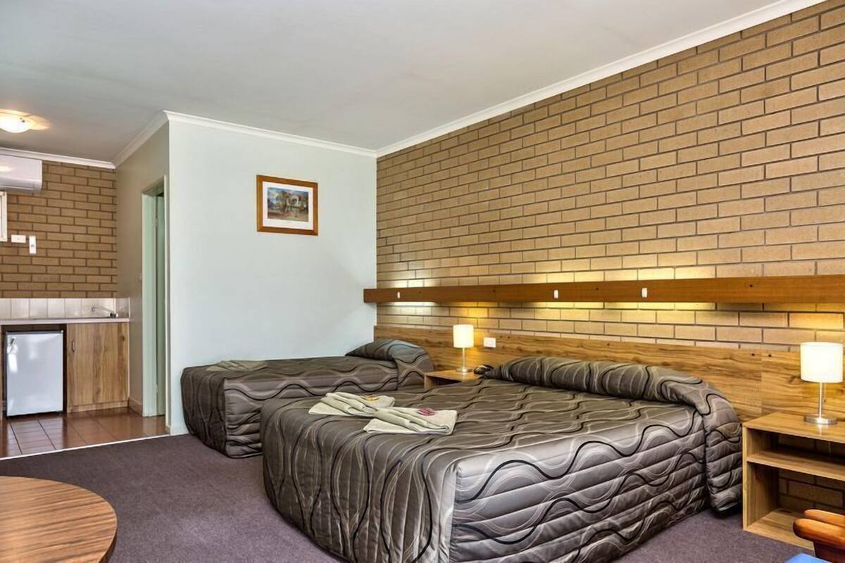 A hotel room with a brick-patterned wall, a wooden headboard, two beds with dark bedding, a small table, and a kitchenette.