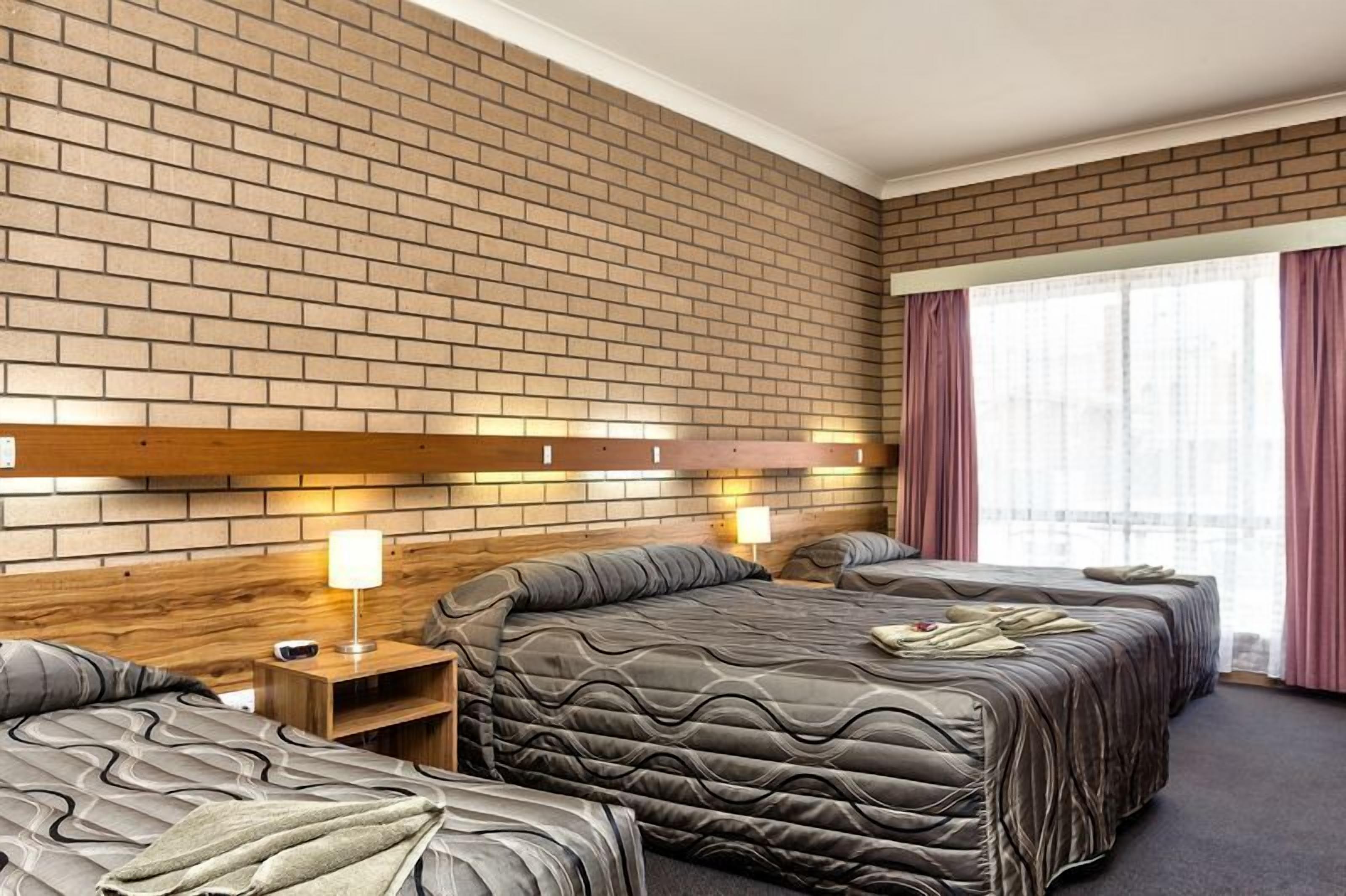 A hotel room with a brick-patterned wall, a wooden headboard, two beds with grey bedding, a nightstand with a lamp, and a large window with curtains.