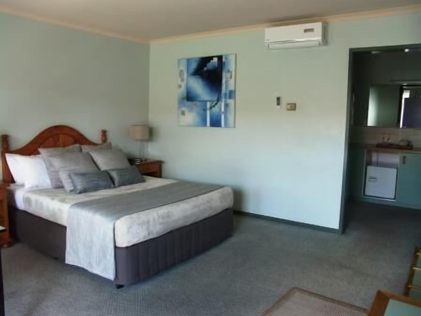 A bedroom with a bed, a nightstand, a lamp, a painting on the wall, and a kitchenette area.