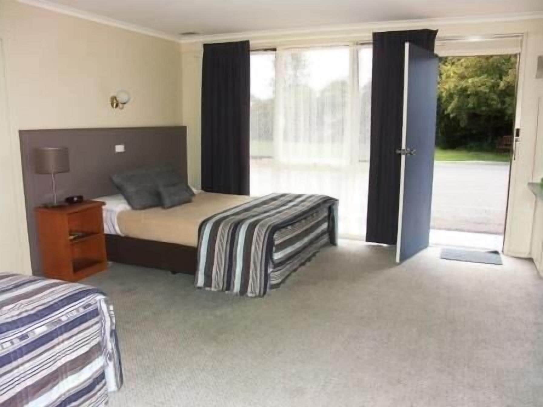 A hotel room with a bed, a nightstand, a lamp, and a view of the outside.