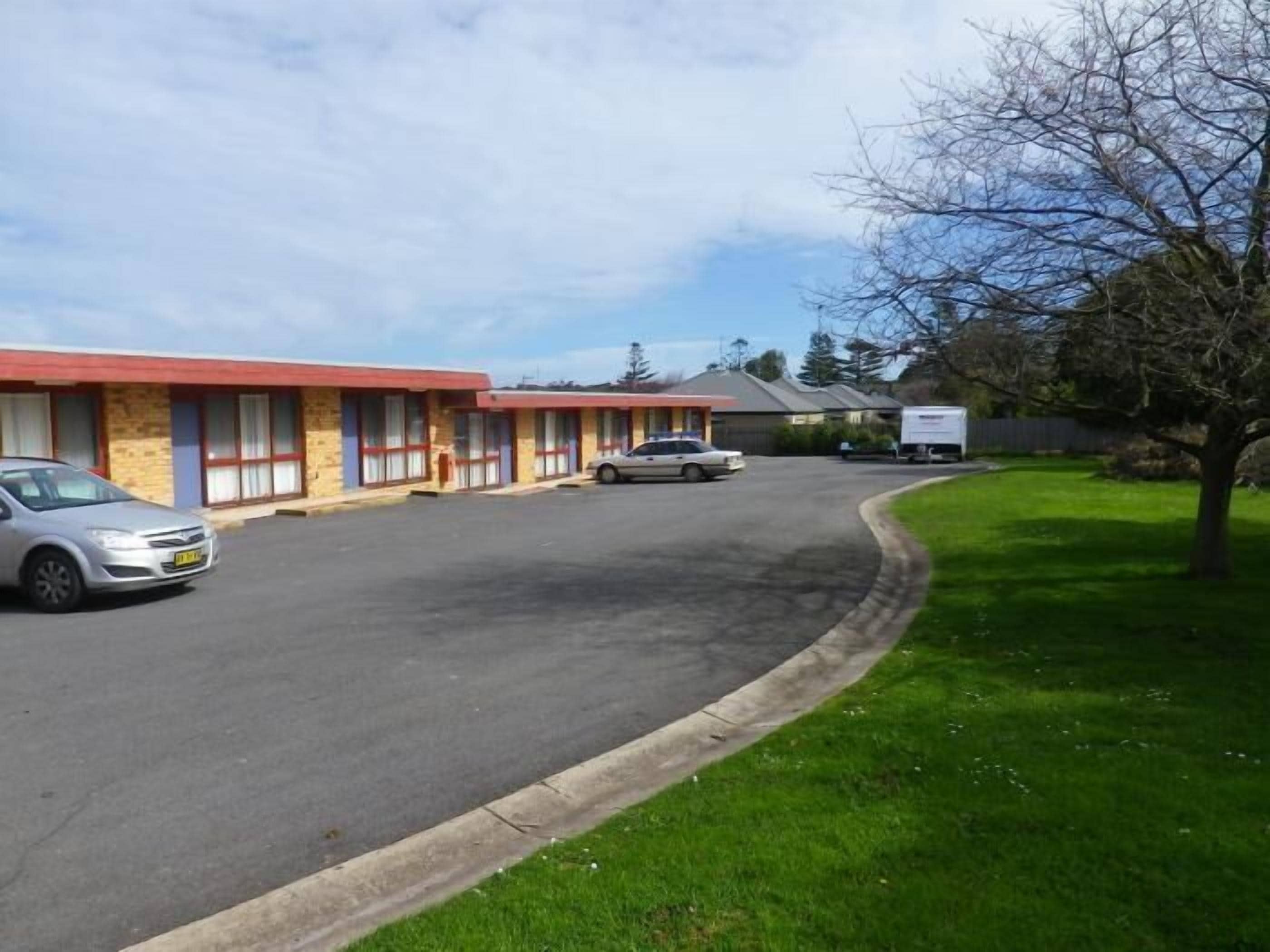 Un motel con una fila de habitaciones, un estacionamiento y un área verde con un árbol.