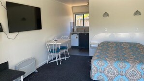 Ein kleines Hotelzimmer mit einem Bett, einem Schreibtisch mit Stuhl, einem Fernseher und einer Küchenzeile im Hintergrund.