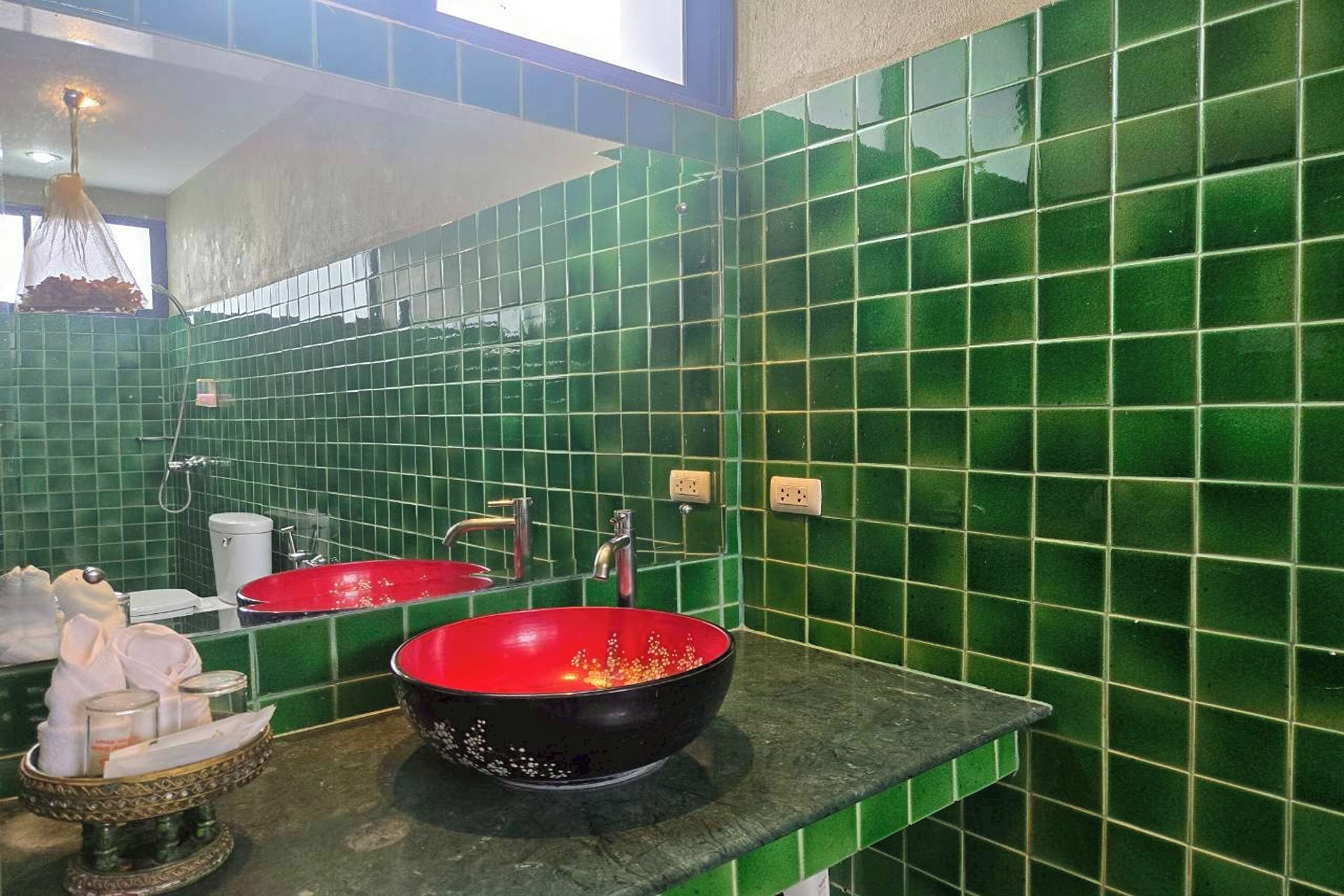 Un baño con paredes cubiertas de azulejos verdes, dos lavabos, un espejo y una lámpara colgante.