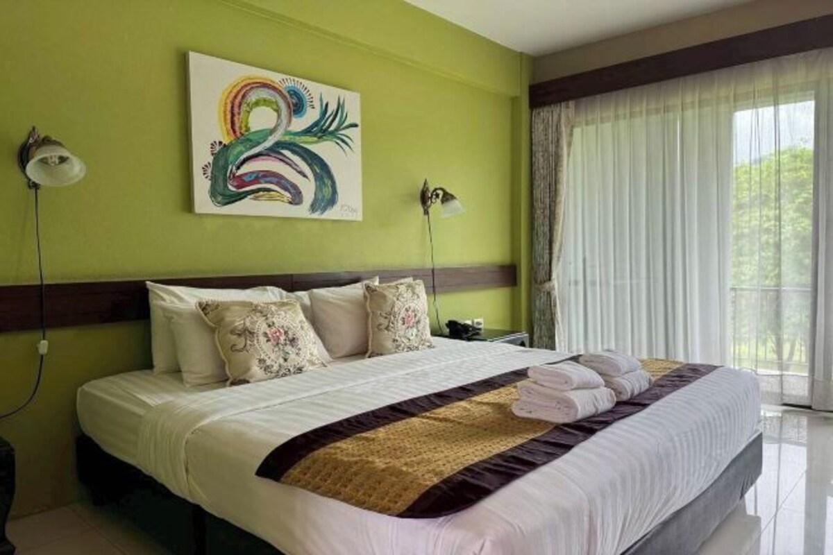 A hotel room with a large bed, a colorful painting on the wall, and a view of greenery through the window.