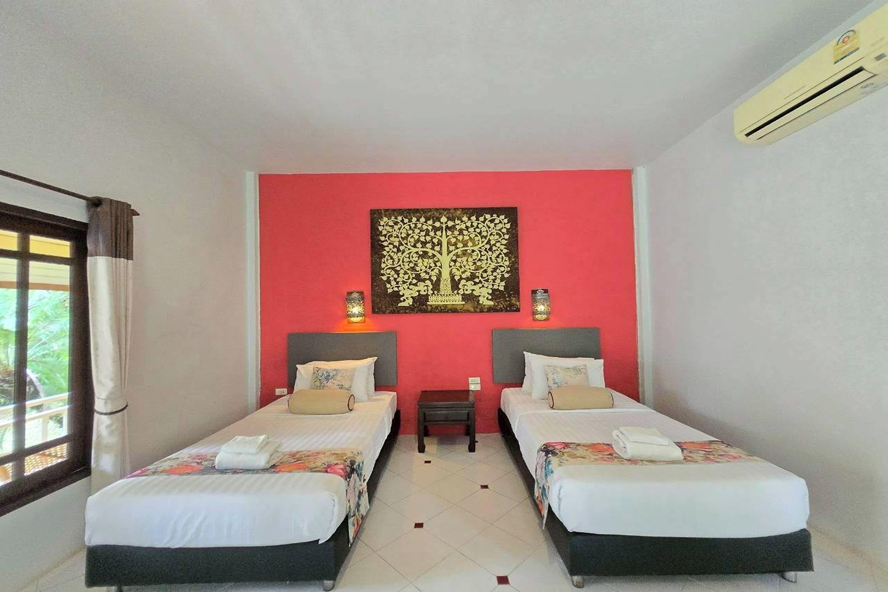 Habitación de hotel con dos camas, una pared de acento roja, un cuadro decorativo de un árbol y un equipo de aire acondicionado.
