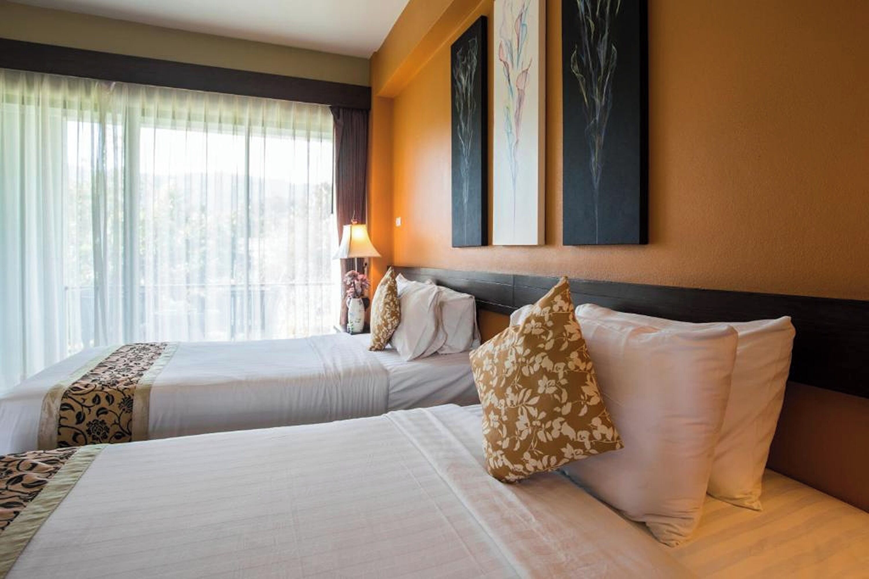A hotel room with two beds, each with white linens and floral-patterned pillows. There are three framed artworks on the wall above the beds.