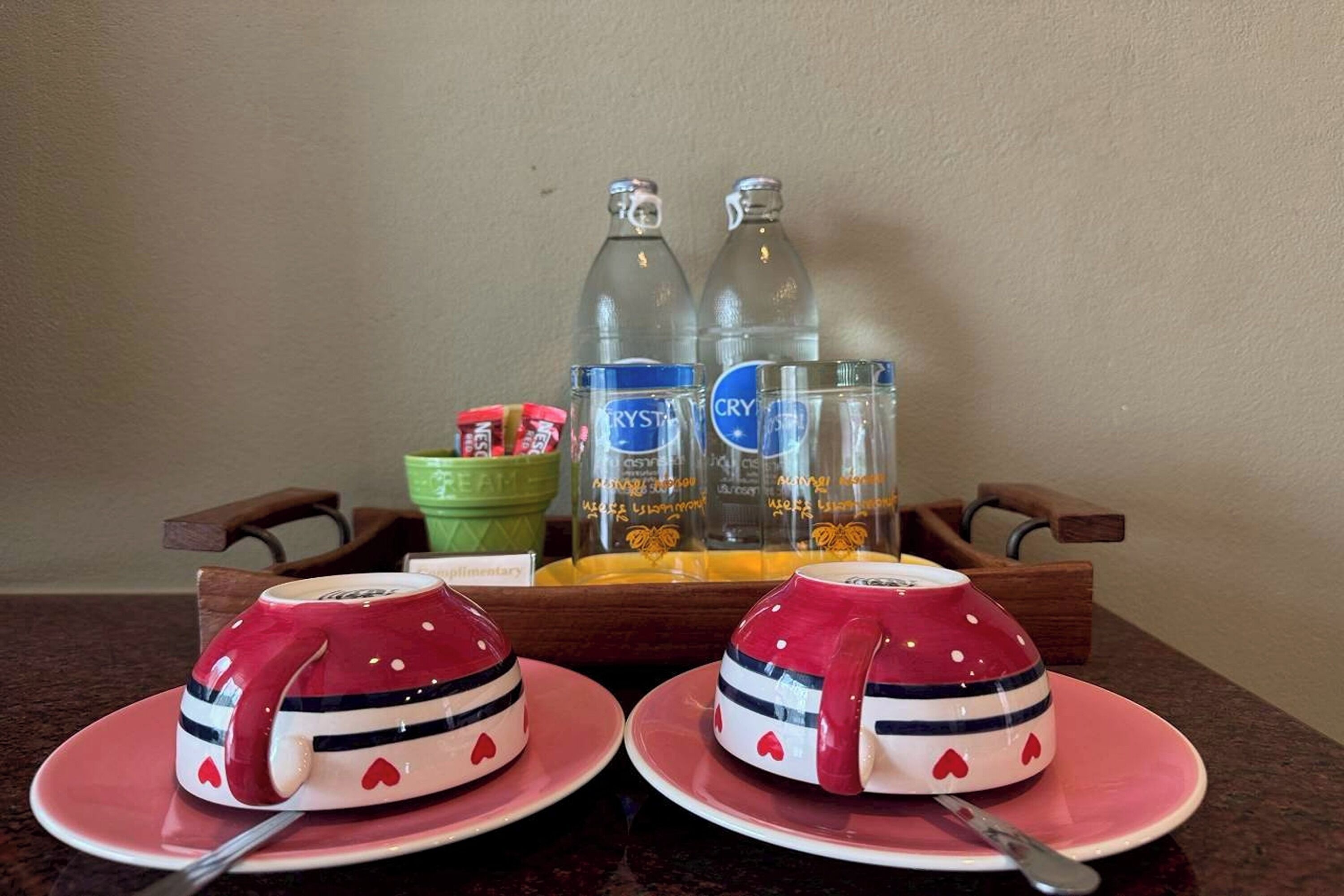A tray with two red cups with heart designs, two clear bottles, a green bowl, and a small container with red and white items.