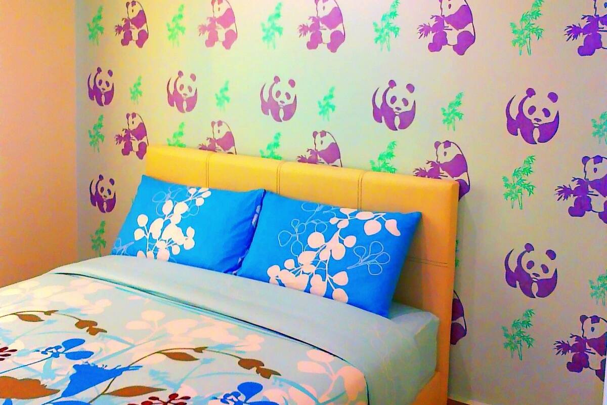 Une chambre à coucher avec un lit, une couvre-lit à motifs floraux bleus et blancs, et un mur décoré d’autocollants représentant des pandas et du bambou.