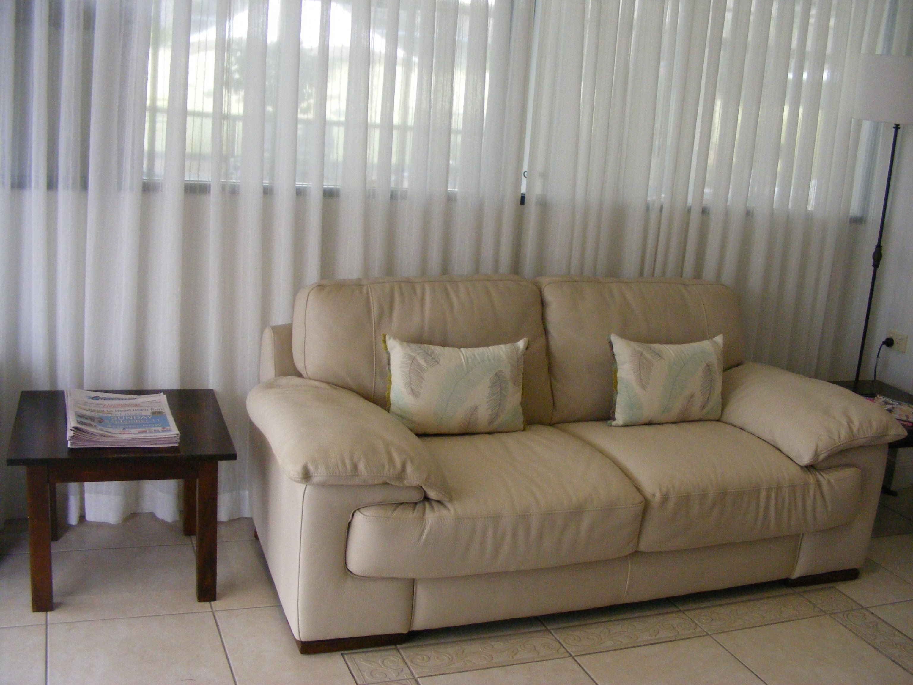 Ein beiges Sofa mit zwei Kissen, ein kleiner Holztisch mit einer Zeitung und ein Fenster mit durchsichtigen Vorhängen.
