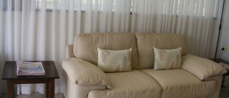 Ein beiges Sofa mit zwei Kissen, ein kleiner Holztisch mit einer Zeitung und ein Fenster mit durchsichtigen Vorhängen.
