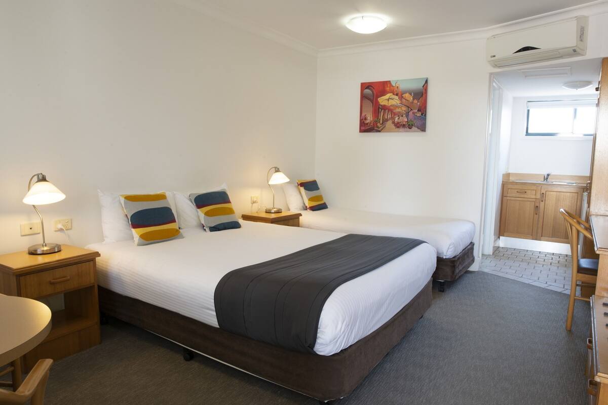 A hotel room with two beds, a desk, a chair, a lamp, a painting on the wall, and a bathroom visible through an open door.