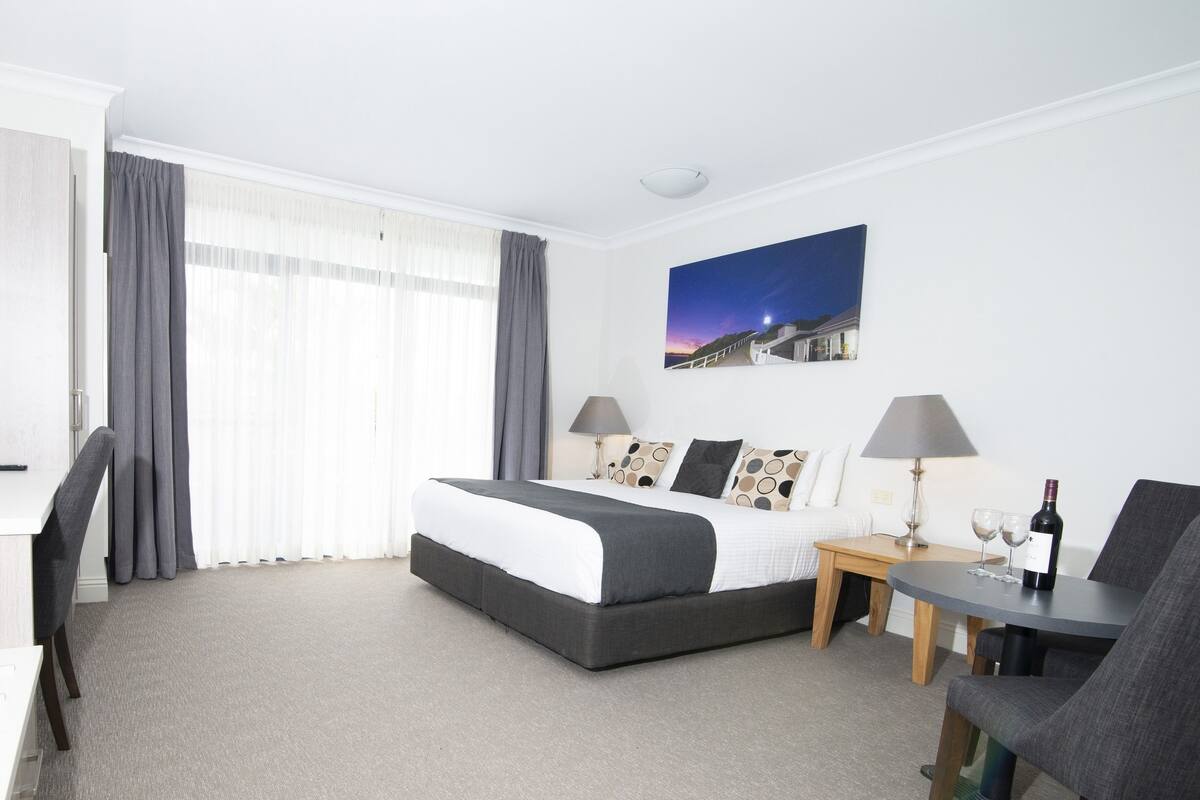 A hotel room with a bed, two bedside tables, a chair, a desk, and a painting on the wall.