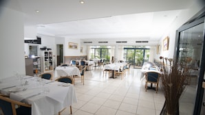 Un vaste espace dédié à la salle à manger, avec plusieurs tables et chaises, un meuble à vin et une vue sur l’extérieur grâce à de grandes fenêtres.