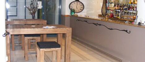 Una zona de bar con mesas y sillas de madera, un reloj en la pared y estantes con botellas.