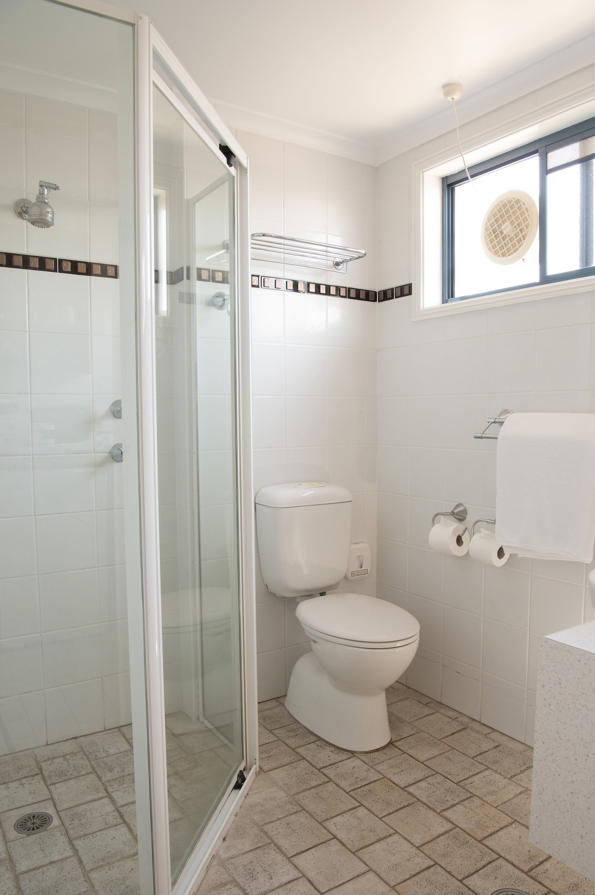Een badkamer met een douche met glazen wand, toilet en raam met plafondventilator.