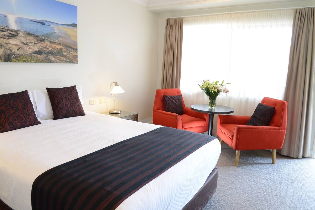 A hotel room with a large bed, two red armchairs, a small round table with a vase of flowers, and a painting of a beach scene on the wall.