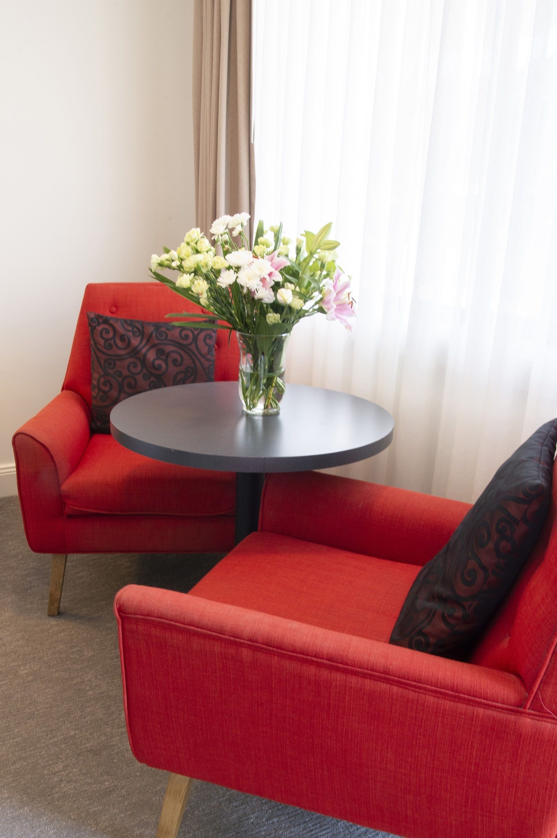 A red sofa with a black patterned pillow, a round table with a vase of flowers, and a window with sheer curtains.
