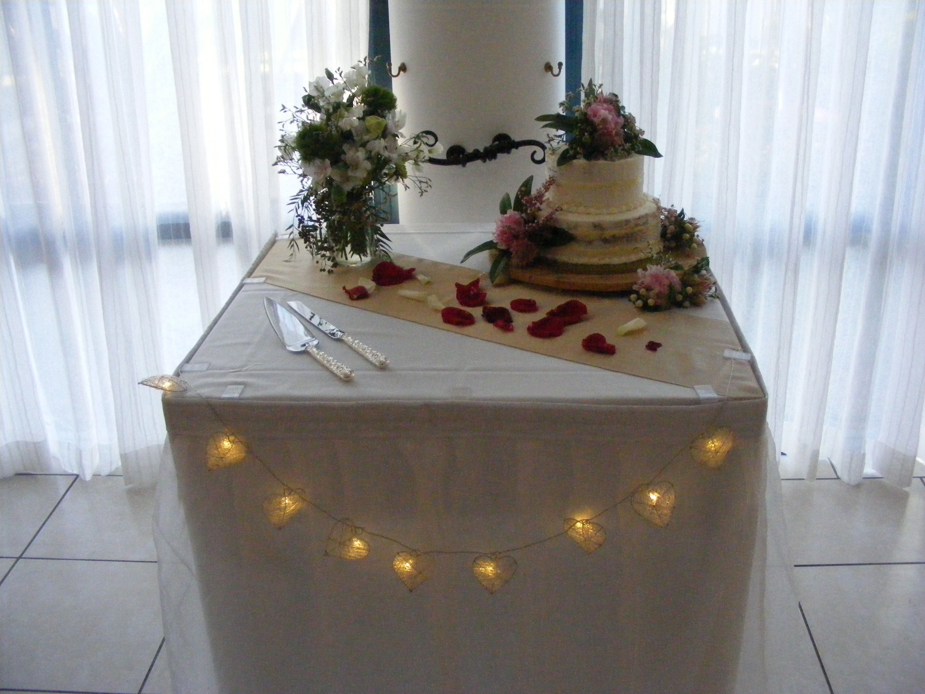 A wedding cake on a table with a white tablecloth, surrounded by flowers and rose petals.