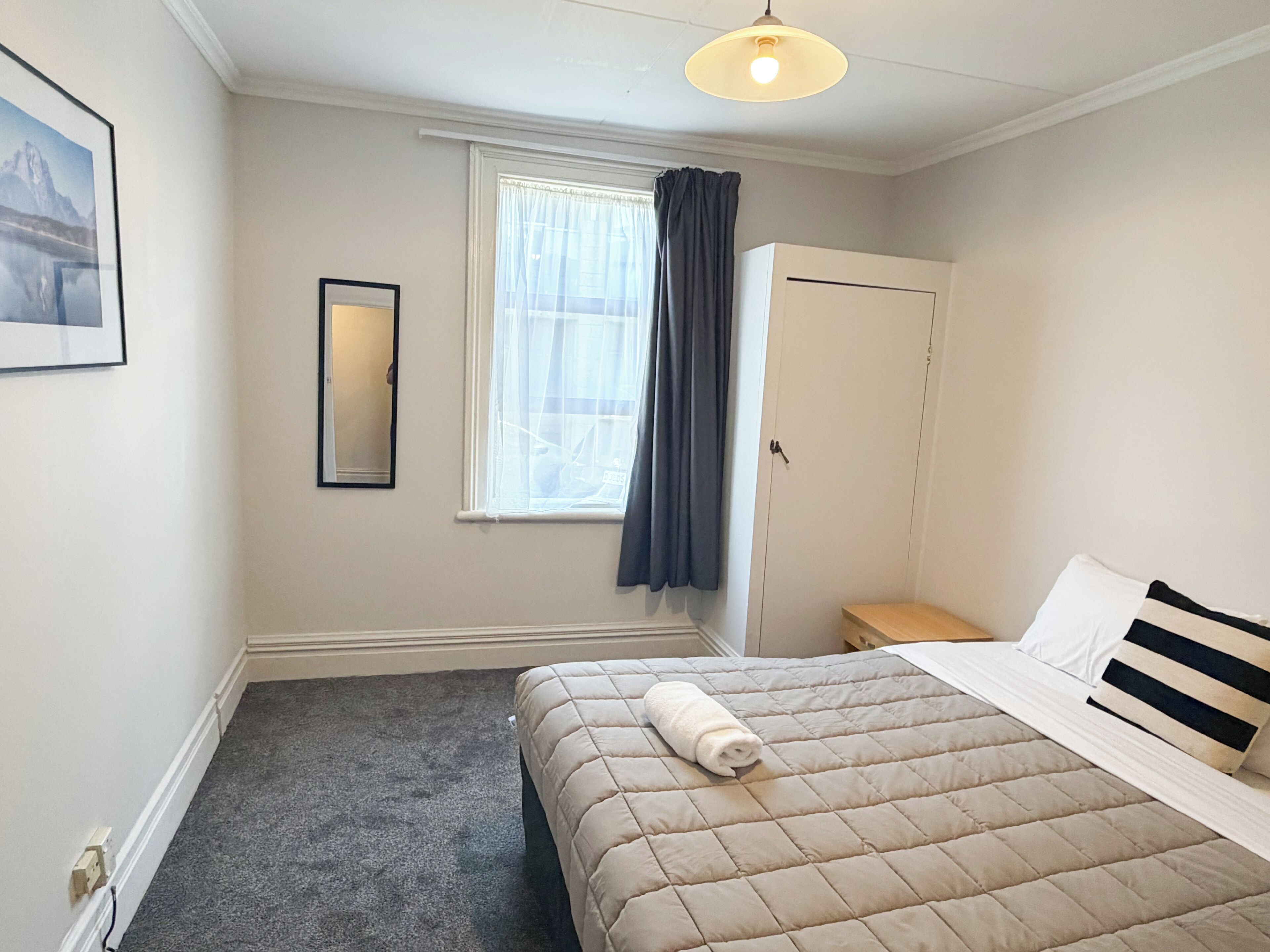 Un dormitorio con cama, armario, ventana con cortinas, espejo y lámpara.