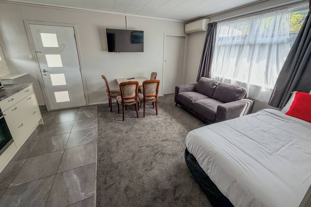 A hotel room with a bed, a sofa, a TV mounted on the wall, a desk with a chair, and a door with frosted glass panels.