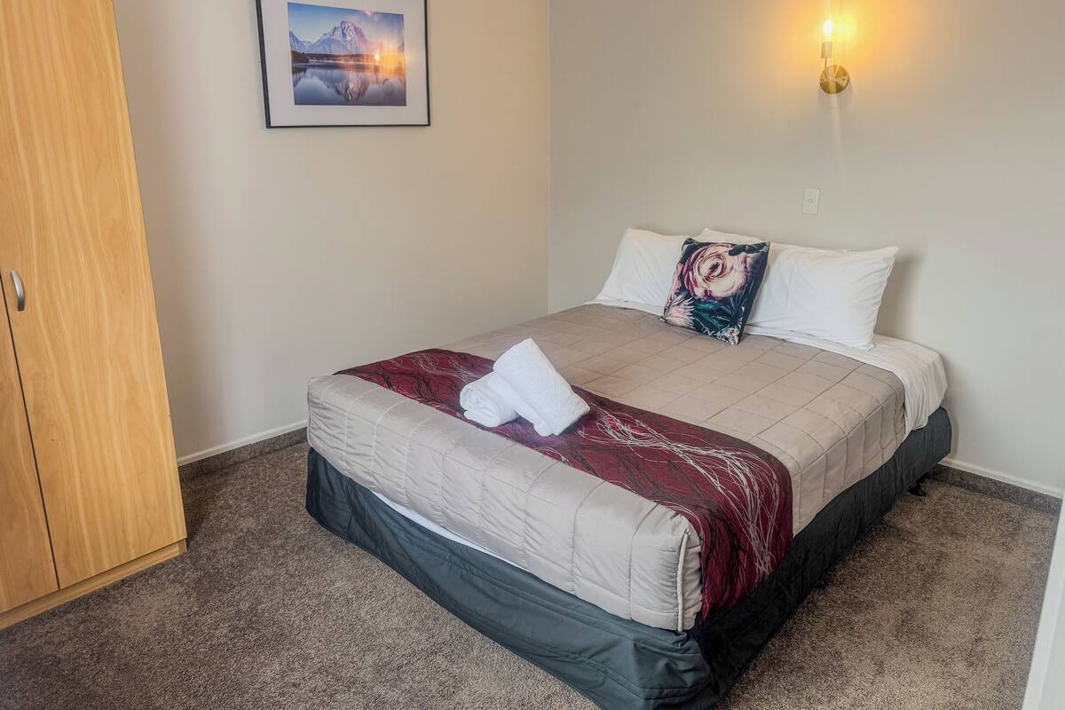 A neatly made bed with a red and white patterned bedspread, a single white pillow, and a decorative pillow with a floral design. A framed picture of a mountain landscape hangs on the wall above the bed.