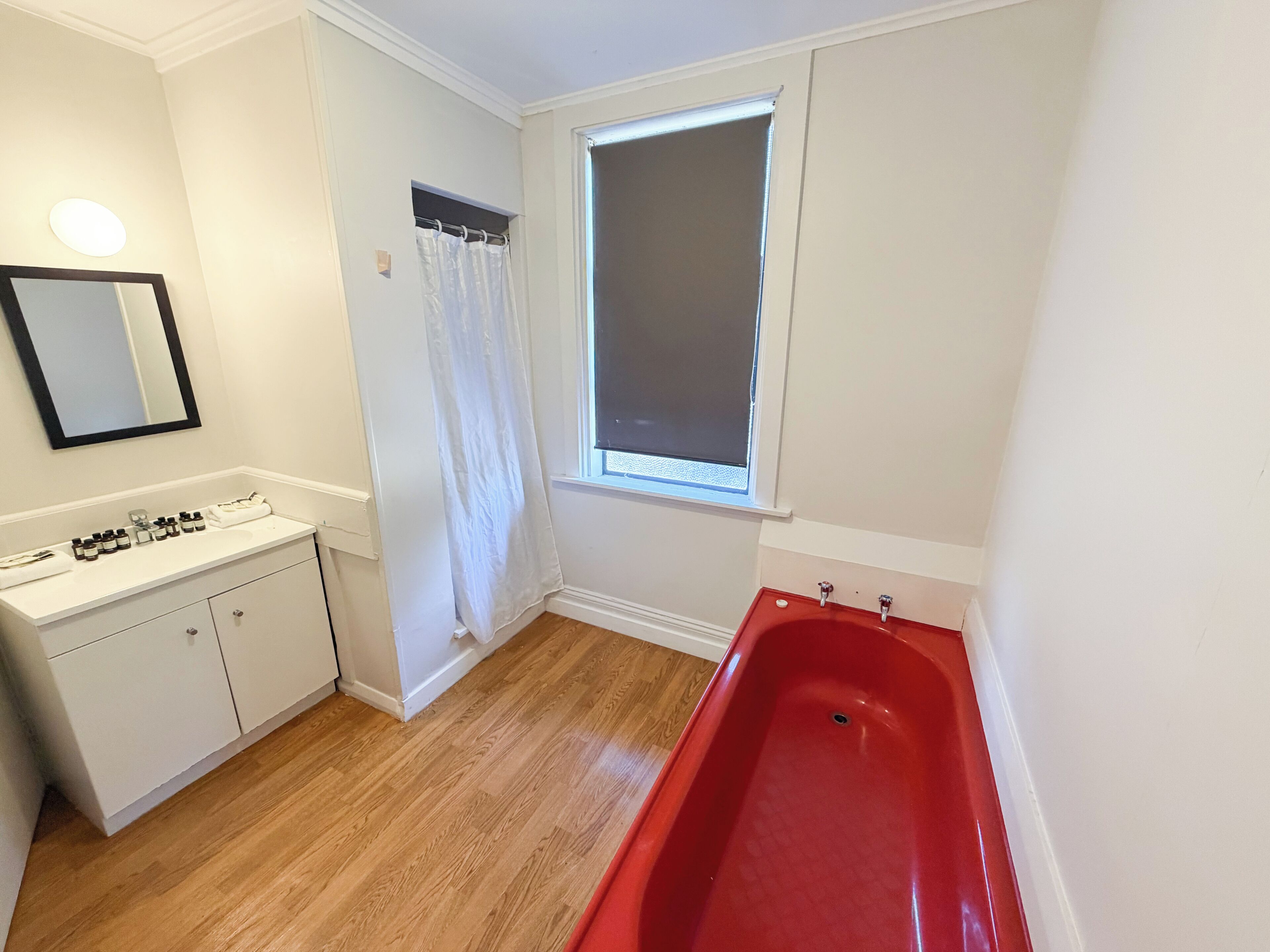 Une salle de bain avec un meuble-lavabo blanc, une baignoire rouge, une fenêtre avec un store et un miroir au mur.