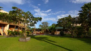 A hotel courtyard with a lawn, trees, and a building with a staircase and outdoor seating. - Rapopo Plantation Resort (Kokopo)