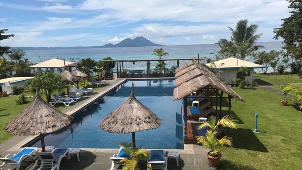 A pool area with lounge chairs and thatched umbrellas, surrounded by greenery and a view of the ocean. - Rapopo Plantation Resort (Kokopo)