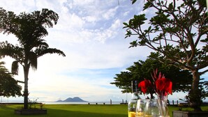 A table with a bottle and a glass of yellow liquid, surrounded by flowers, set outdoors with a view of a grassy field and a distant mountain. - Rapopo Plantation Resort (Kokopo)