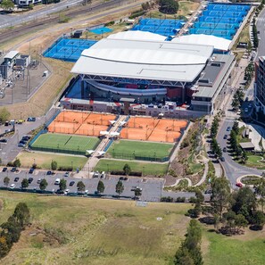 An aerial view of a sports complex with multiple tennis courts, a large stadium, and surrounding parking areas.