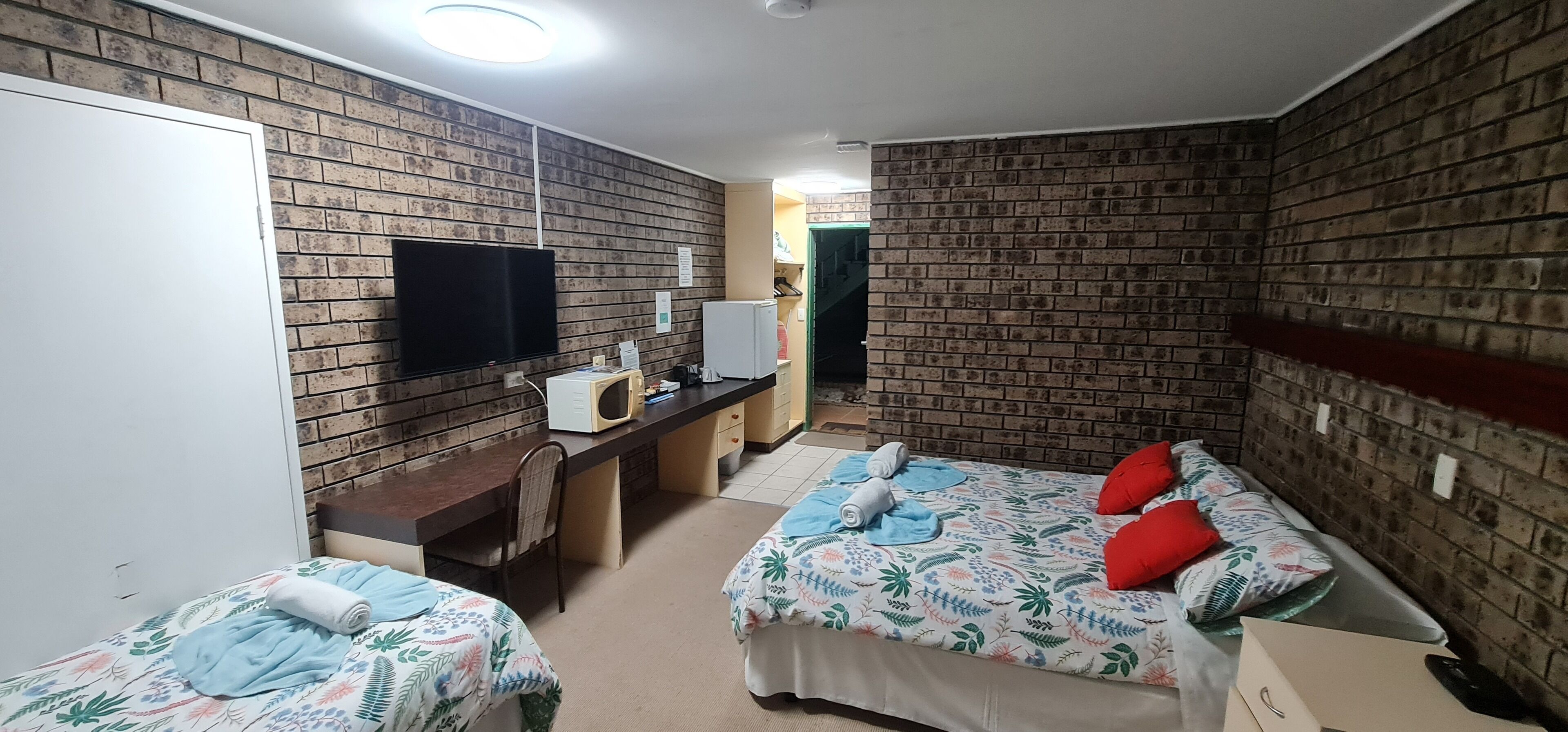 A hotel room with brick-patterned walls, a flat-screen TV, a desk with a microwave, two beds with floral bedding, and a wardrobe.