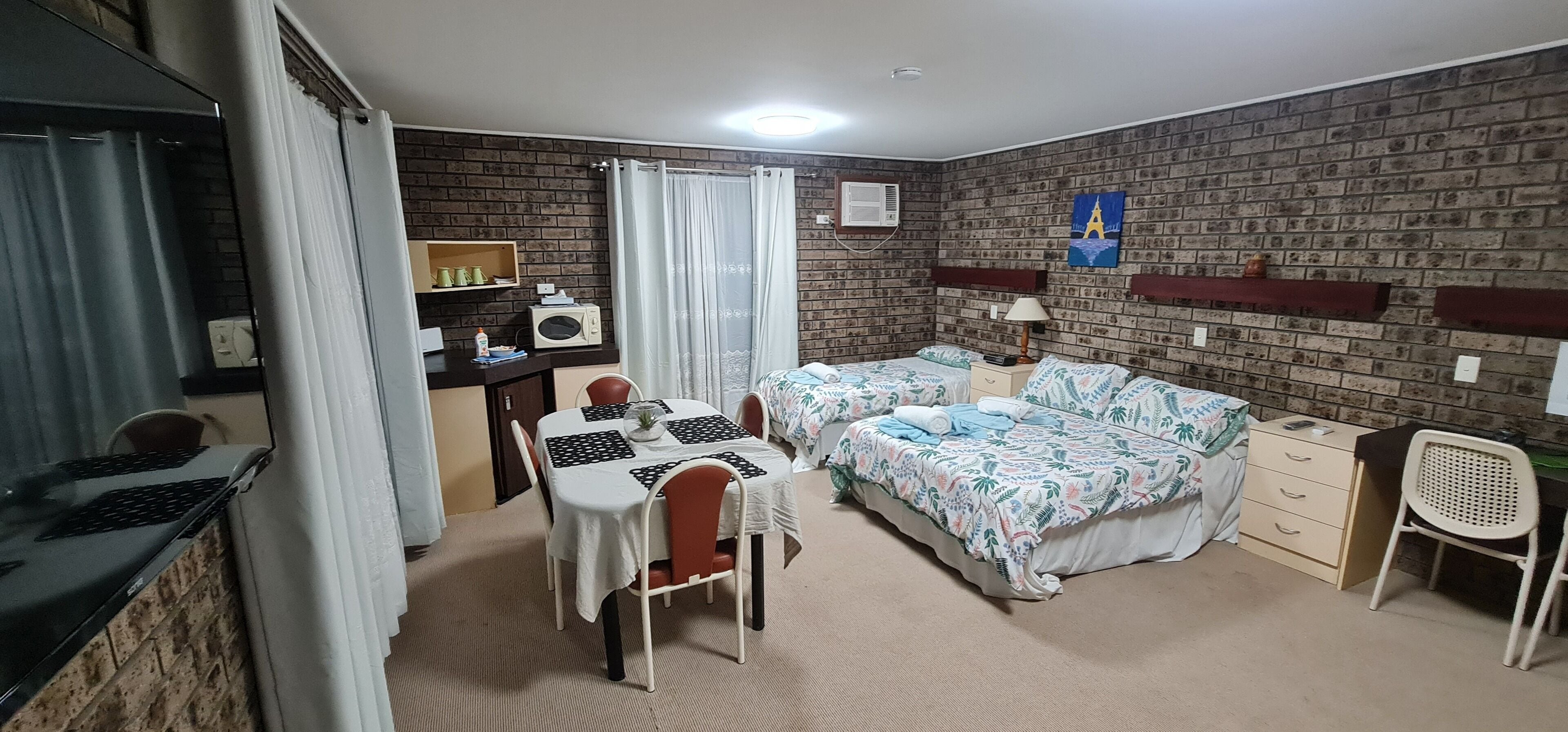 A room with a brick wall, two beds with floral bedding, a desk with a laptop, and a small table with chairs.