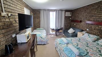 A hotel room with brick walls, a flat-screen TV, a desk with a kettle, two beds with floral bedding, a sofa, and a small table with chairs.