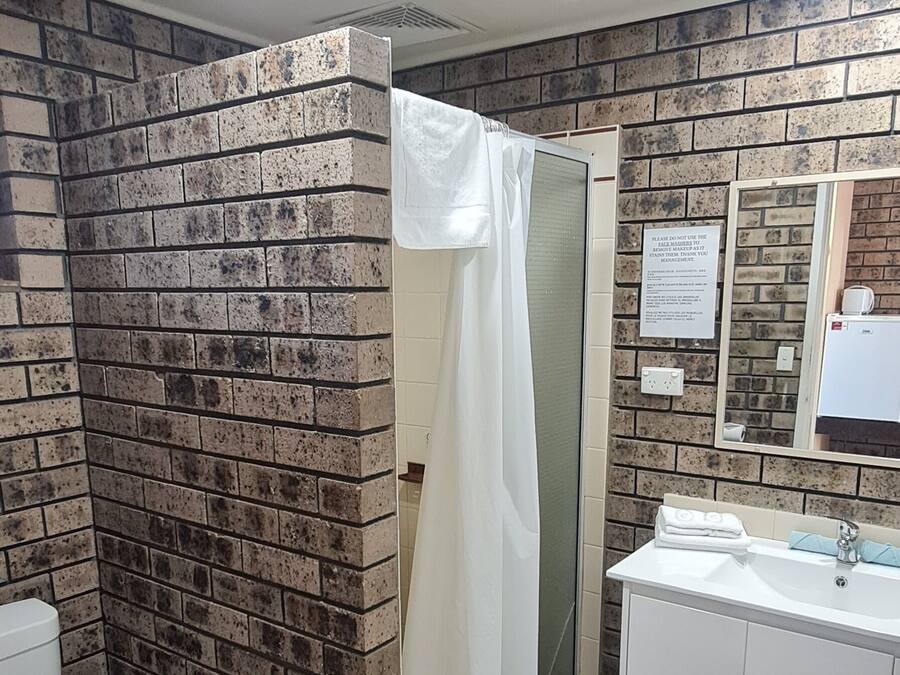 Une salle de bain avec un mur en briques, des toilettes blanches, une douche avec un rideau blanc, un lavabo avec un miroir et un tapis blanc sur le sol.